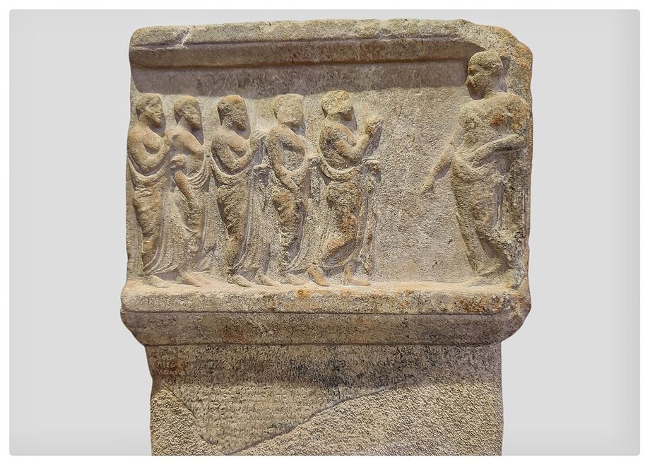 Upper part of an Artemis Brauronia's stele with a scene and a catalogue of dedications to Artemis Brauronia. Around 420 B.C.  On the right, Artemis wearing a chiton and himation is leaning against a pilaster holding a phiale. Five men wearing himation advance towards the goddess with respect. These may perhaps be interpreted as the board of 'Hieropoioi' or the 'the Treasurers of the Other Gods', operating in the sanctuary in weighing and/or counting the sacred property before its transfer to the Acropolis. In the inventory of the sacred treasure of Artemis the sacred money of Apollo is also mentioned. This was possibly part of the Allies' Treasure, which had been transferred from Delos to Athens (453 B.C.).  The inscription in the field of the relief: ΦΙΛΟΜ ΦΙΛΟΜΕΛΟΣ was carved in later years. It apparently praises the known family of Philomeledai from Marathon who took part in the transfer of the goddess' property from the Brauron sanctuary to the Athens Acropolis.