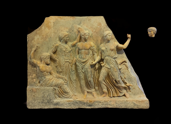 The subject of the scene on the relief of the Gods is Iphigeneia's arrival at Brauron and the establishment of the sanctuary according to the tradition mentioned by Pausanias. The first figure on the left is a bearded man, possibly Zeus, seated on a low rock and holding with his right hand a cane or a mace, which was painted. A mature female figure, Leto, follows. She wears a peplos covering part of her head and holds a painted mace. Apollo is standing next to her possibly with a branch of laurel in his hand. He just like  Zeus and Leto are facing right. The female figure wearing the attic peplos is Artemis. She is running to the left but she is facing right as rest of the figures. The holes on her tight fists imply that she was holding bronze objects, possibly a torch and a bow.  At the right part of the scene, which is missing, the two siblings, Orestes and Iphigeneia were possibly depicted on a chariot drawn by two deer. Orestes had just pulled the bridle of the chariot and the deer sat up on their back feet while Iphigeneia stood holding the goddess's effigy in her hands. The head at the right of the remaining relief is identified with Iphigeneia.  According to other interpretations, Artemis arrives at Brauron with a chariot drawn by deer while the figure wearing a peplos is identified with Iphigeneia,  who either holds two torches or the bridles of the chariot. The male figure at the beginning of the scene has also been attributed to Brauron, the eponymous hero of Brauron.