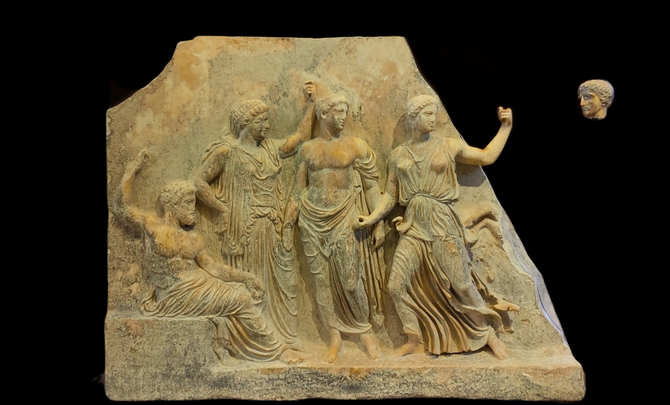 The subject of the scene on the relief of the Gods is Iphigeneia's arrival at Brauron and the establishment of the sanctuary according to the tradition mentioned by Pausanias. The first figure on the left is a bearded man, possibly Zeus, seated on a low rock and holding with his right hand a cane or a mace, which was painted. A mature female figure, Leto, follows. She wears a peplos covering part of her head and holds a painted mace. Apollo is standing next to her possibly with a branch of laurel in his hand. He just like  Zeus and Leto are facing right. The female figure wearing the attic peplos is Artemis. She is running to the left but she is facing right as rest of the figures. The holes on her tight fists imply that she was holding bronze objects, possibly a torch and a bow.  At the right part of the scene, which is missing, the two siblings, Orestes and Iphigeneia were possibly depicted on a chariot drawn by two deer. Orestes had just pulled the bridle of the chariot and the deer sat up on their back feet while Iphigeneia stood holding the goddess's effigy in her hands. The head at the right of the remaining relief is identified with Iphigeneia.  According to other interpretations, Artemis arrives at Brauron with a chariot drawn by deer while the figure wearing a peplos is identified with Iphigeneia,  who either holds two torches or the bridles of the chariot. The male figure at the beginning of the scene has also been attributed to Brauron, the eponymous hero of Brauron.